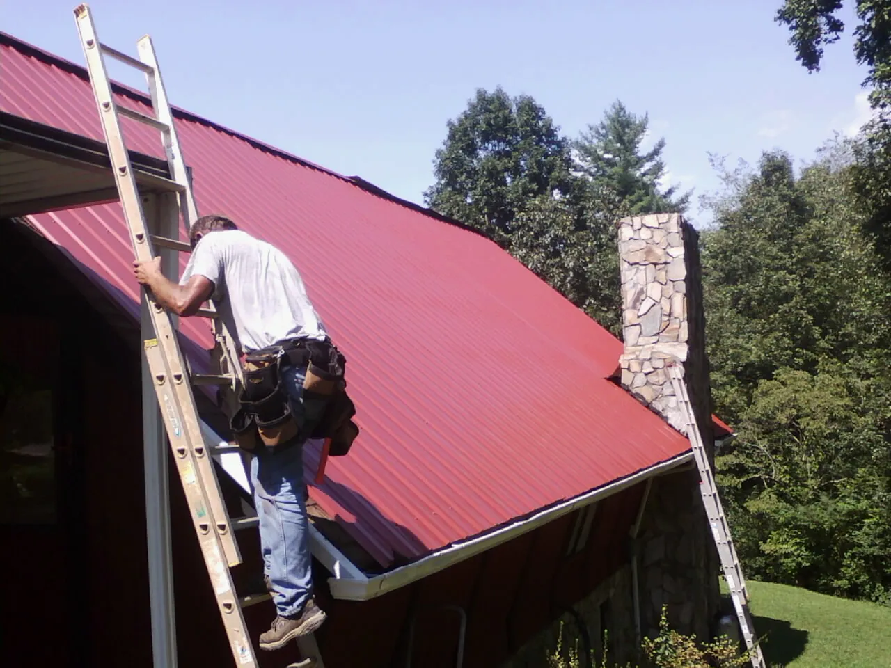 Roofer on a ladder beside a red metal roof and stone chimney — Soffit & Fascia Repair by Roofing Brothers in Hiddenite